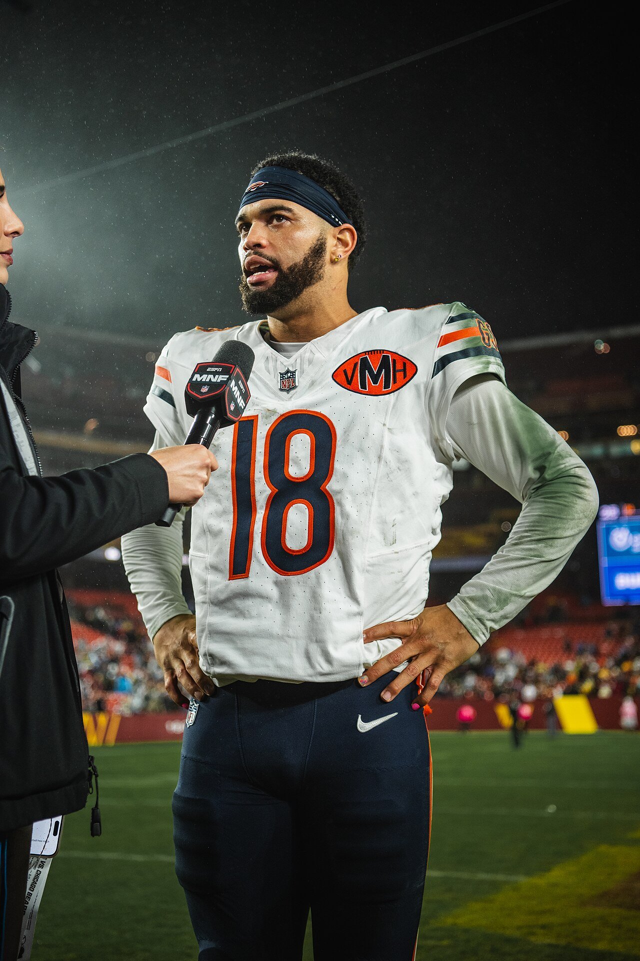 Bears quarterback Caleb Williams in his white number 18 jersey speaking into a microphone on the field after a night game